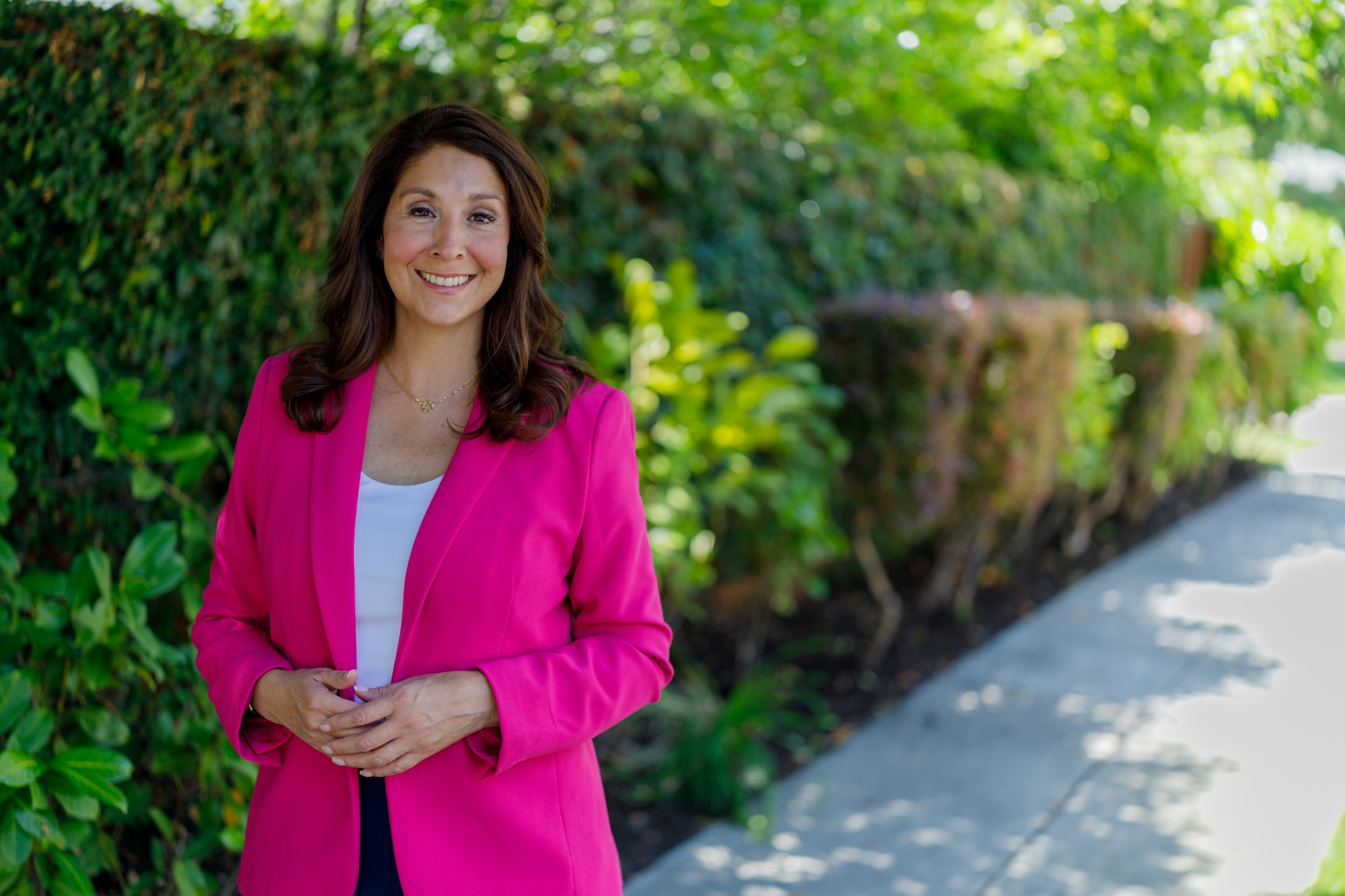 Melissa Hernandez smiling in a pink blazer on a tree-lined sidewalk